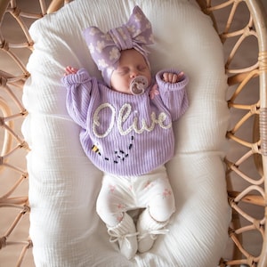 May include: A baby wearing a lavender sweater with the name "Olive" embroidered on the front, paired with floral leggings and a matching bow headband. The sweater features a bee design. The baby is in a wicker bassinet.
