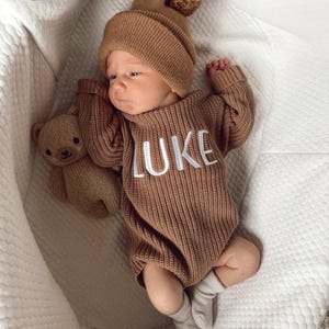 May include: A newborn baby wearing a brown knit romper with the name "LUKE" embroidered in white. The baby is also wearing a matching knit hat with a pom-pom and is lying next to a small teddy bear. The baby is wearing gray socks.