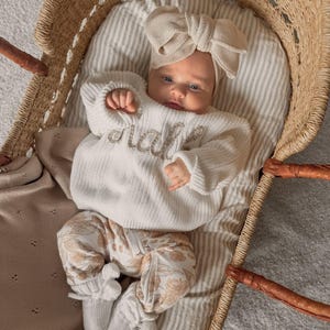 May include: A baby in a cream knitted sweater with the name "malia" embroidered on the front, floral trousers, and a matching bow headband. The baby lies in a wicker bassinet with a striped blanket.