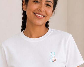 May include: White t-shirt with a small, abstract design in red and blue on the chest. The shirt has short sleeves and a crew neck. The model is wearing beige pants. The background includes a framed artwork, a plant, and decorative items.