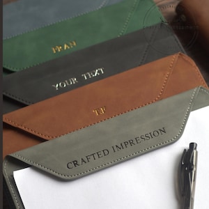 May include: A selection of personalised leather document holders in grey, brown, black, and green. Each holder has gold lettering with initials or text. A black pen rests on a white sheet of paper.