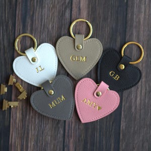May include: Five heart-shaped keyrings in various colours: white, grey, black, pink, and taupe. Each keyring has gold lettering and a gold ring. The keyrings are personalised with initials and names. They are on a dark wood surface.