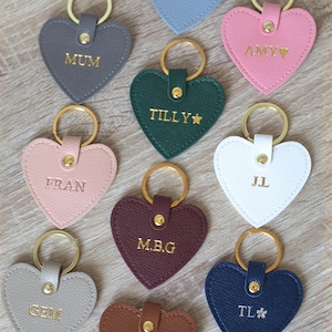 Può includere: Una collezione di portachiavi a forma di cuore in vari colori, tra cui grigio, rosa, verde, bianco e blu. Ogni portachiavi presenta un anello dorato e nomi o iniziali personalizzati in lettere dorate. Disposti su una superficie di legno chiaro.