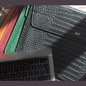 May include: Leather laptop sleeves in a range of colours: black, green, red, and orange. The black sleeve is embossed with the gold initials "M.S". A laptop keyboard is partially visible in the foreground.