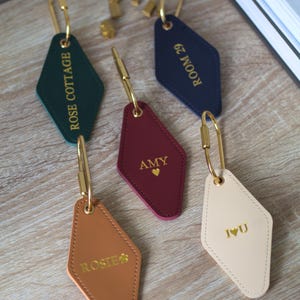 May include: A selection of diamond-shaped keyrings in shades of green, navy, burgundy, tan, and cream. Each keyring features gold text and a gold clasp. The text includes names and phrases such as "Rose Cottage" and "Room 29".