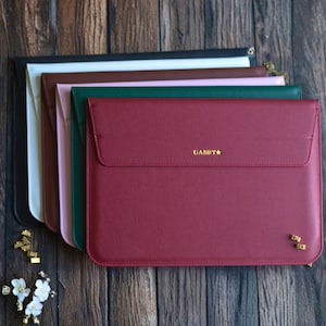 Może przedstawiać: Stos kolorowych etui na laptopy w kolorach czarnym, białym, brązowym, różowym, zielonym i bordowym. Bordowe etui ma złoty napis "Gabby". Etui ułożone są na drewnianej powierzchni.