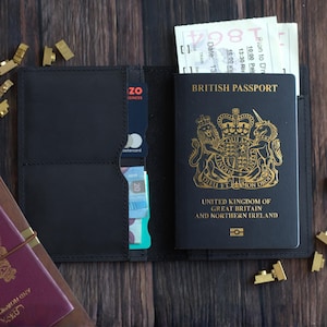 K&ouml;nnte beinhalten: Ge&ouml;ffnete schwarze Leder-Passh&uuml;lle mit britischem Reisepass, Tickets und einer Kreditkarte. Der Reisepass ist schwarz mit goldfarbenen Buchstaben und einem k&ouml;niglichen Wappen.