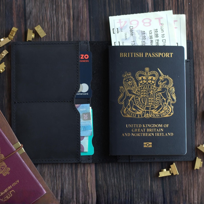 K&ouml;nnte beinhalten: Ge&ouml;ffnete schwarze Leder-Passh&uuml;lle mit britischem Reisepass, Tickets und einer Kreditkarte. Der Reisepass ist schwarz mit goldfarbenen Buchstaben und einem k&ouml;niglichen Wappen.