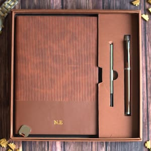 May include: A brown stationery gift set in a rectangular box containing a notebook with a textured, ribbed upper cover and a smooth lower section featuring the gold embossed initials N.E., accompanied by a matching brown and silver metal pen.