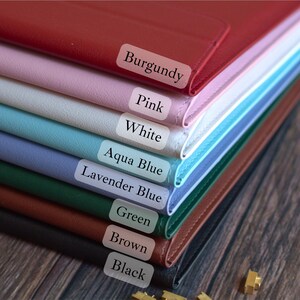 May include: A stack of leather goods in various colors, including burgundy, pink, white, aqua blue, lavender blue, green, brown, and black. Each item is labeled with its color name. The items are stacked on top of each other.