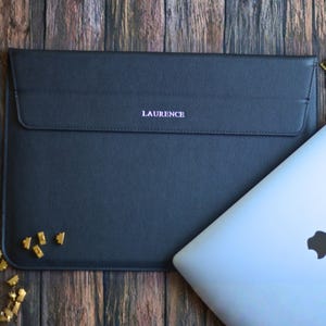 May include: A black laptop sleeve with the name "LAURENCE" printed in a light pink font. The sleeve has a flap closure. A silver laptop is partially visible. The sleeve is made of a smooth material.
