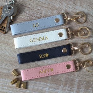 May include: Four rectangular keychains in light blue, white, black, and pink with gold-tone hardware. Each features a gold-stamped name or initials: J.C, GEMMA, RS with a flower icon, and AMY with a heart icon. Each has a gold snap button.