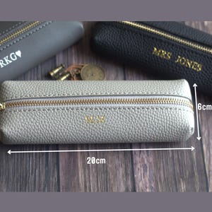 May include: A light gray pencil case with a gold zipper and the initials "M.M." in gold. The case is 20 cm long and 6 cm high. Other cases in gray and black are visible in the background.