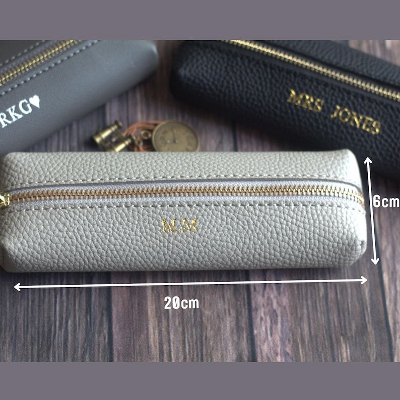 May include: A light gray pencil case with a gold zipper and the initials "M.M." in gold. The case is 20 cm long and 6 cm high. Other cases in gray and black are visible in the background.