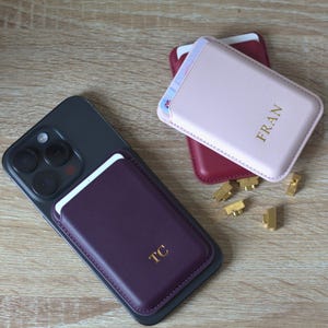 May include: A dark purple magnetic card holder attached to a black smartphone, featuring the gold embossed initials TC. Nearby, a light pink magnetic card holder with the gold embossed name FRAN, and a dark red card holder, with small gold metal pieces.