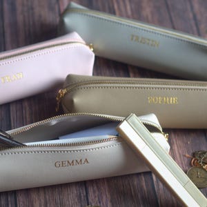 Op de afbeelding: Een verzameling gepersonaliseerde etuis in verschillende neutrale kleuren, waaronder roze, beige en grijs. Elke etui heeft een gouden rits en een in goudreliëf aangebrachte naam. Eén etui is open en toont pennen en potloden.