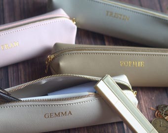 May include: A collection of personalized pencil cases in various neutral colors, including pink, beige, and gray. Each case has a gold zipper and is embossed with a name in gold lettering. One case is open, revealing pens and pencils.