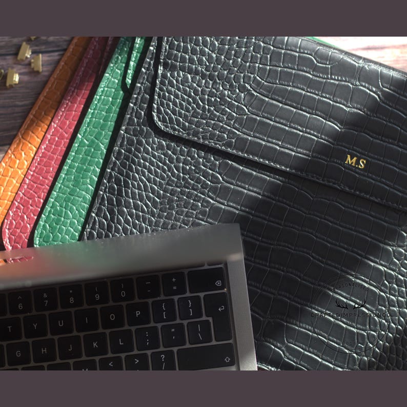 May include: A collection of leather laptop sleeves in various colors, including black, green, red, and orange. The black sleeve features the initials "M.S" in gold. A laptop keyboard is visible in the foreground.