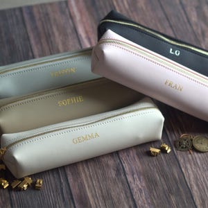 May include: A collection of personalized pencil cases in various colors, including black, pink, beige, and gray. Each case features a gold zipper and a name in gold lettering. The cases are arranged on a wooden surface.