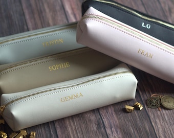 May include: A collection of personalized pencil cases in various colors, including black, pink, beige, and gray. Each case features a gold zipper and a name in gold lettering. The cases are arranged on a wooden surface.