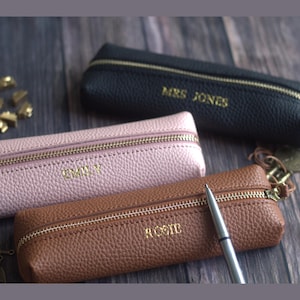 May include: Three personalized pencil cases in black, pink, and brown leather, each with a gold zipper and a name embossed in gold lettering. A silver pen rests on the brown case. The cases are on a dark wood surface.