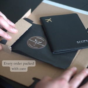 May include: Black leather passport holder with a gold airplane icon and the name "SCOTT" in white. The passport holder is packaged in a black paper sleeve with a brown cardboard box. Text on the box reads "Every order packed with care."