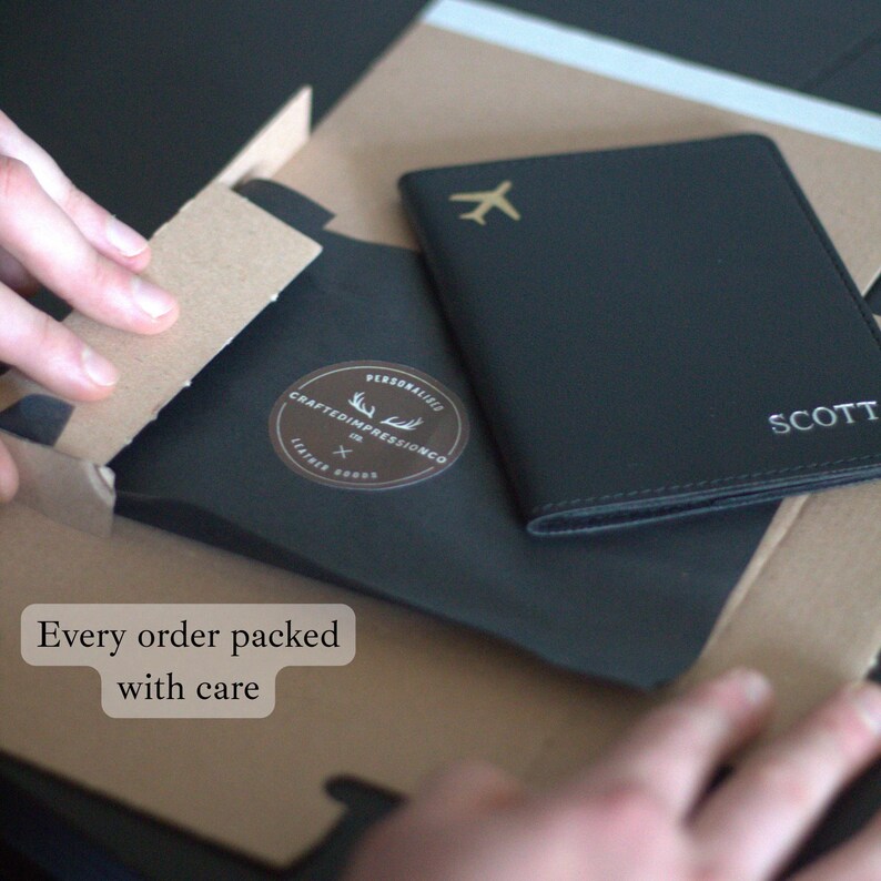 May include: Black leather passport holder with a gold airplane icon and the name "SCOTT" in white. The passport holder is packaged in a black paper sleeve with a brown cardboard box. Text on the box reads "Every order packed with care."
