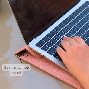 May include: A laptop computer rests on a pink, textured laptop stand. The stand is built-in and supports the laptop at an angle. The keyboard is visible, and a hand is typing on the keys. The text "Built in Laptop Stand" is also visible.