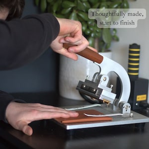 May include: A silver and brown leather crafting machine with a wooden handle is used to press a piece of brown leather. The text "Thoughtfully made, start to finish" is in the upper right corner. The machine is on a metal surface.