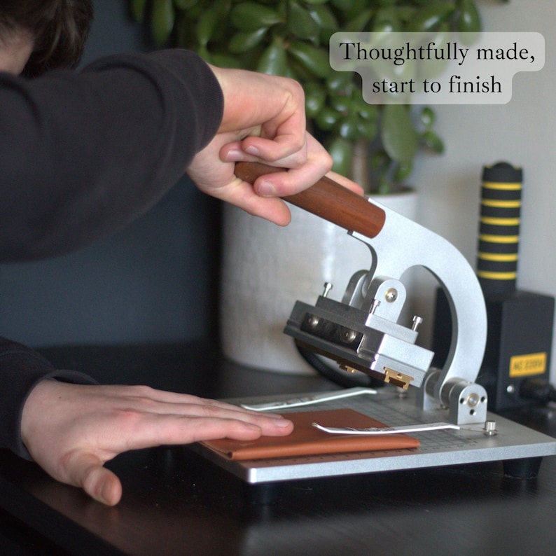 May include: A silver and brown leather crafting machine with a wooden handle is used to press a piece of brown leather. The text "Thoughtfully made, start to finish" is in the upper right corner. The machine is on a metal surface.