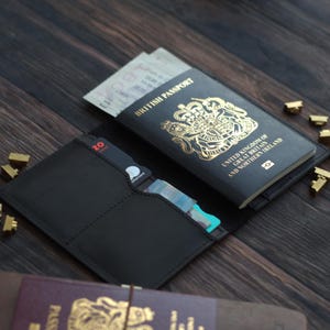 K&ouml;nnte beinhalten: Schwarzer Leder-Reisepasshalter mit einem britischen Reisepass und Karten. Der Reisepass ist schwarz mit goldfarbenen Buchstaben und einem k&ouml;niglichen Wappen. Der Halter ist offen und zeigt das Innere. Goldfarbene Metallakzente sind zu sehen.