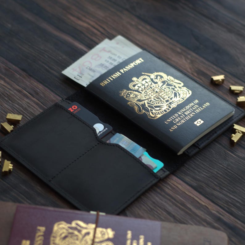 K&ouml;nnte beinhalten: Schwarzer Leder-Reisepasshalter mit einem britischen Reisepass und Karten. Der Reisepass ist schwarz mit goldfarbenen Buchstaben und einem k&ouml;niglichen Wappen. Der Halter ist offen und zeigt das Innere. Goldfarbene Metallakzente sind zu sehen.
