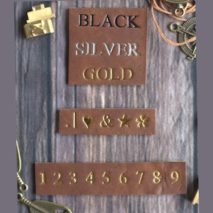 Può includere: Primo piano di quadrati di pelle marrone con testo e simboli in rilievo. Il quadrato superiore recita "BLACK", "SILVER" e "GOLD". Sotto ci sono simboli e i numeri da 1 a 9. Accenti metallici decorativi sullo sfondo.