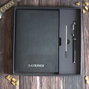 Può includere: Un set regalo nero contenente un taccuino rettangolare con texture a coste e il nome LAURENCE impresso in argento sulla copertina. Il set include una penna a sfera nera abbinata e un singolo refill d'inchiostro argentato in una scatola.