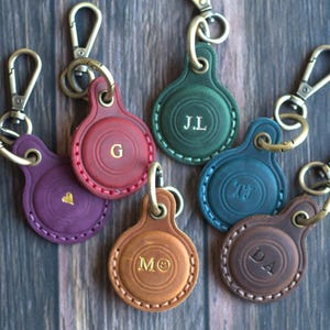 Könnte beinhalten: Eine Auswahl an Leder-Schlüsselanhängern in verschiedenen Farben, darunter Lila, Rot, Grün, Blau, Braun und Beige. Jeder Schlüsselanhänger hat ein rundes Design mit einem goldenen Initial oder Herz. Sie haben goldfarbene Metallverschlüsse.