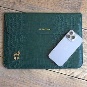 May include: A dark green, textured laptop sleeve with a flap closure. The sleeve has the text "M.SMITH" in gold lettering, along with a small gold emblem. A silver smartphone rests on the sleeve, which is placed on a wooden surface.