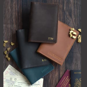 K&ouml;nnte beinhalten: Eine Auswahl an personalisierten Leder-Reisepassh&uuml;llen in verschiedenen Farben: Dunkelbraun, Hellbraun, Schwarz und T&uuml;rkis. Jede H&uuml;lle ist mit einem Namen oder Initialen in Gold gepr&auml;gt. Reiseaccessoires.