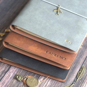 May include: Three leather journals in grey, brown, and black are stacked. The top journal has a gold airplane charm and the initials "R.G." embossed. The middle journal is embossed with the name "LAURENCE".