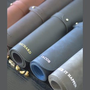 May include: Rolled leather desk mats in shades of grey, blue, and brown. Each mat is personalised with a name in gold or white lettering. The mats are secured with a strap and button closure.