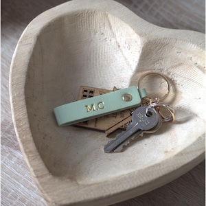 Può includere: Un portachiavi in pelle verde chiaro con le iniziali M.C. impresse in oro e un anello in metallo argentato. È dotato di un piccolo ciondolo in legno a forma di casa con testo visibile e una chiave in metallo argentato con bordo seghettato.