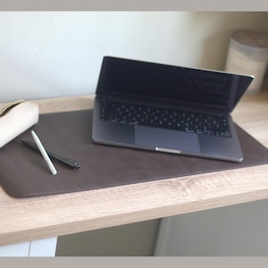 May include: A brown leather desk mat with a laptop, pens, and a pencil case on a light wood desk. The laptop is open, displaying the keyboard. A white pencil and a black pen are on the mat. The pencil case is off-white.