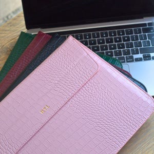 Può includere: Una collezione di custodie per laptop in vari colori, tra cui rosa, verde, bordeaux e nero. Le custodie presentano un motivo strutturato simile alla pelle di coccodrillo e una chiusura a patta. La custodia rosa ha le iniziali "HTL" in oro.