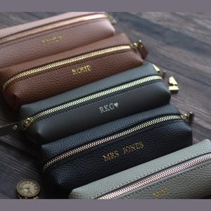 May include: A collection of personalized pencil cases in various colors, including tan, brown, gray, and black. Each case features a gold zipper and is embossed with a name or initials. The cases are arranged on a dark wooden surface.