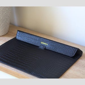 May include: Black faux-leather laptop sleeve with a crocodile skin texture. The flap is embossed with the name "GEMMA" in gold lettering. The sleeve is open, revealing the interior.