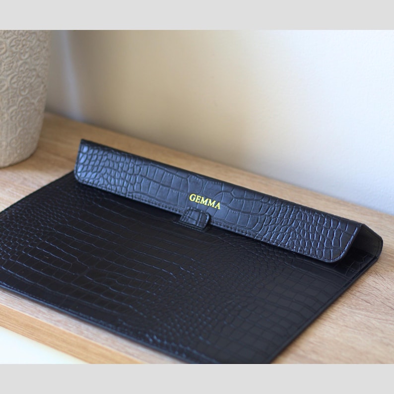 May include: Black faux-leather laptop sleeve with a crocodile skin texture. The flap is embossed with the name "GEMMA" in gold lettering. The sleeve is open, revealing the interior.