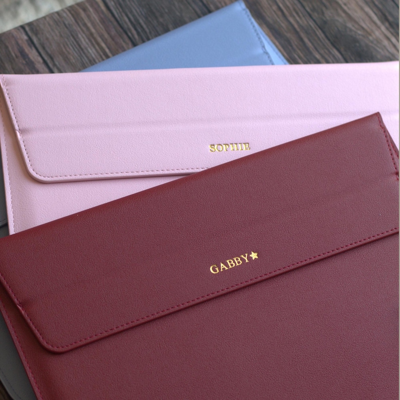 May include: A close-up of three personalized laptop sleeves in shades of burgundy, pink, and light blue. Each sleeve has a flap closure and is embossed with a name in gold lettering. The sleeves are made of a smooth, leather-like material.