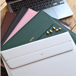 May include: A selection of laptop sleeves in colours such as white, green, pink, black, and brown. Each sleeve is personalised with gold initials or a name. Gold-coloured decorative accents are also visible.