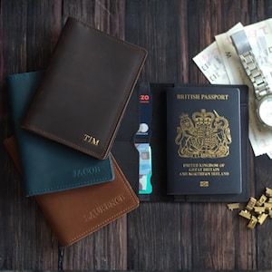 May include: A collection of personalized leather passport holders in various colors, including brown, teal, and tan, with names embossed in gold. A British passport, watch, and travel documents are also visible.