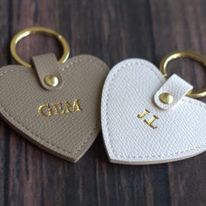 May include: Two heart-shaped keyrings. One is taupe with gold lettering "GEM", the other is white with gold lettering "JL". Both have gold key rings and matching gold hardware.