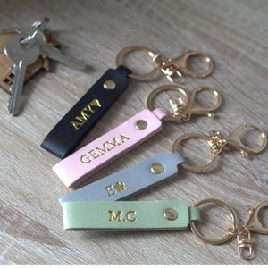 May include: Four rectangular faux leather keychains in black, light pink, grey, and light green. Each features gold-tone hardware, a metal snap, and gold embossed lettering reading AMY, GEMMA, E, and M.C. A set of silver keys is attached to the black keychain.